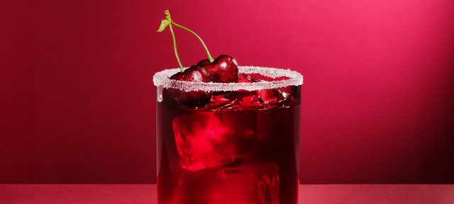 Glas mit ECKES Cherry Sour Cocktail, garniert mit einer Kirsche und Zuckerrand, vor einem roten Hintergrund