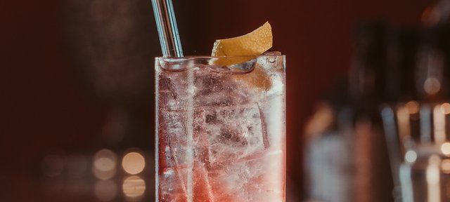 Glas mit ECKES Cherry Highball Cocktail auf Eis, garniert mit einer Orangenzeste und einem Strohhalm, vor einem unscharfen Barhintergrund