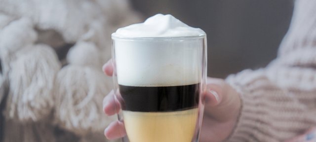 Hand hält ein Glas mit ECKES Calimero Espresso, geschichtet mit Sahne, vor einem gemütlichen Hintergrund.