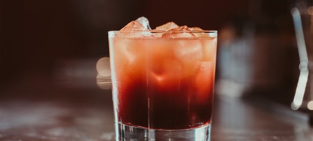 Glas mit ECKES Blood & Sand Cocktail auf Eiswürfeln, vor einem unscharfen, dunklen Hintergrund.