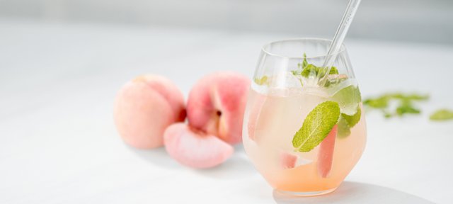 Glas mit ECKES White Peach Cocktail, garniert mit Minze und Pfirsichscheiben, vor einem weißen Hintergrund mit frischen Pfirsichen.