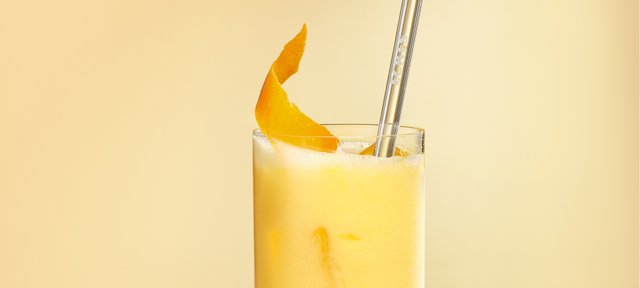 Glas mit ECKES Egg Blond Cocktail, garniert mit einer Zitronenzeste und einem Strohhalm, vor einem gelben Hintergrund.