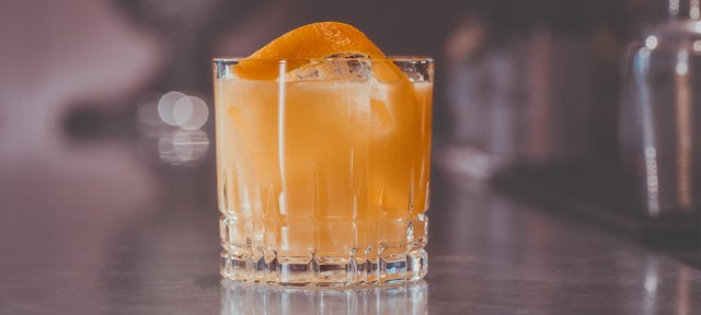 Glas mit ECKES Casablanca Cocktail auf Eis, garniert mit einer Orangenschale, vor einem unscharfen Barhintergrund.