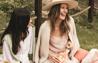 Zwei lachende Frauen sitzen im Freien, eine mit Strohhut und eine Flasche in der Hand, die andere mit einem Cocktailglas.