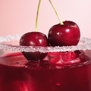 Zwei Kirschen auf einem Glas mit rotem Cocktail und Zuckerrand, vor einem rosa Hintergrund.