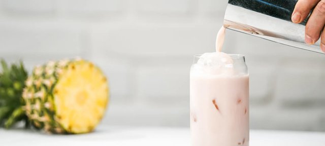 Glas mit ECKES Peach Cola Cocktail, über das gerade Flüssigkeit gegossen wird, vor einem weißen Hintergrund mit einer Ananas.