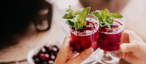  Zwei Personen stoßen mit Gläsern Cherry Smash Cocktail an, garniert mit frischen Minzblättern.