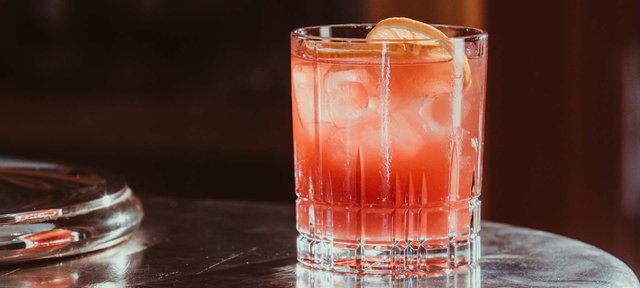 Glas mit ECKES Cherry Mash Sour Cocktail auf Eis, garniert mit einer Orangenscheibe, auf einem dunklen Tisch.