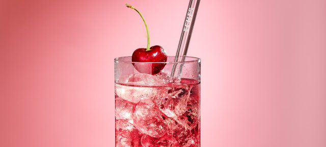 Glas mit ECKES Cherry Tonic Cocktail, garniert mit einer Kirsche und einem Strohhalm, vor einem rosafarbenen Hintergrund.