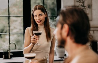 Eine Frau in einem eleganten, cremefarbenen Kleid hält ein Cocktailglas und blickt lächelnd zu einem Mann, der im Vordergrund sitzt.