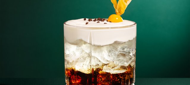 Glas mit ECKES White Russian Cocktail auf Eis, garniert mit Sahne und Orangenschale, vor einem grünen Hintergrund.