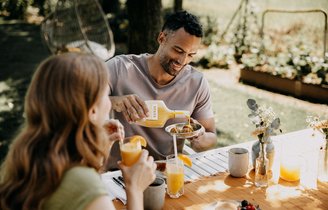 Ein Mann und eine Frau sitzen an einem Tisch im Freien, der Mann gießt Eierlikör in ein Cocktailglas, während die Frau einen Eierlikör-Drink genießt.